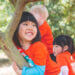 Young girl smiling sitting on and holding onto a tree trunk with other kids.
