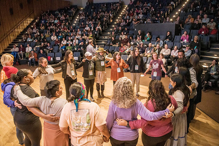Group standing in a circle together in an auditorium with a crowd in the backdrop.