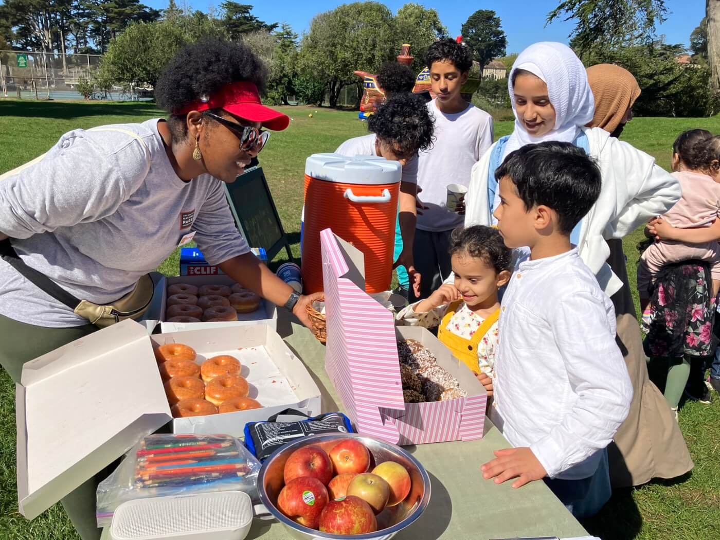 table with assortment of donuts and fruit in the park. Woman is on one side of the table smiling at the children on the other side of the table.