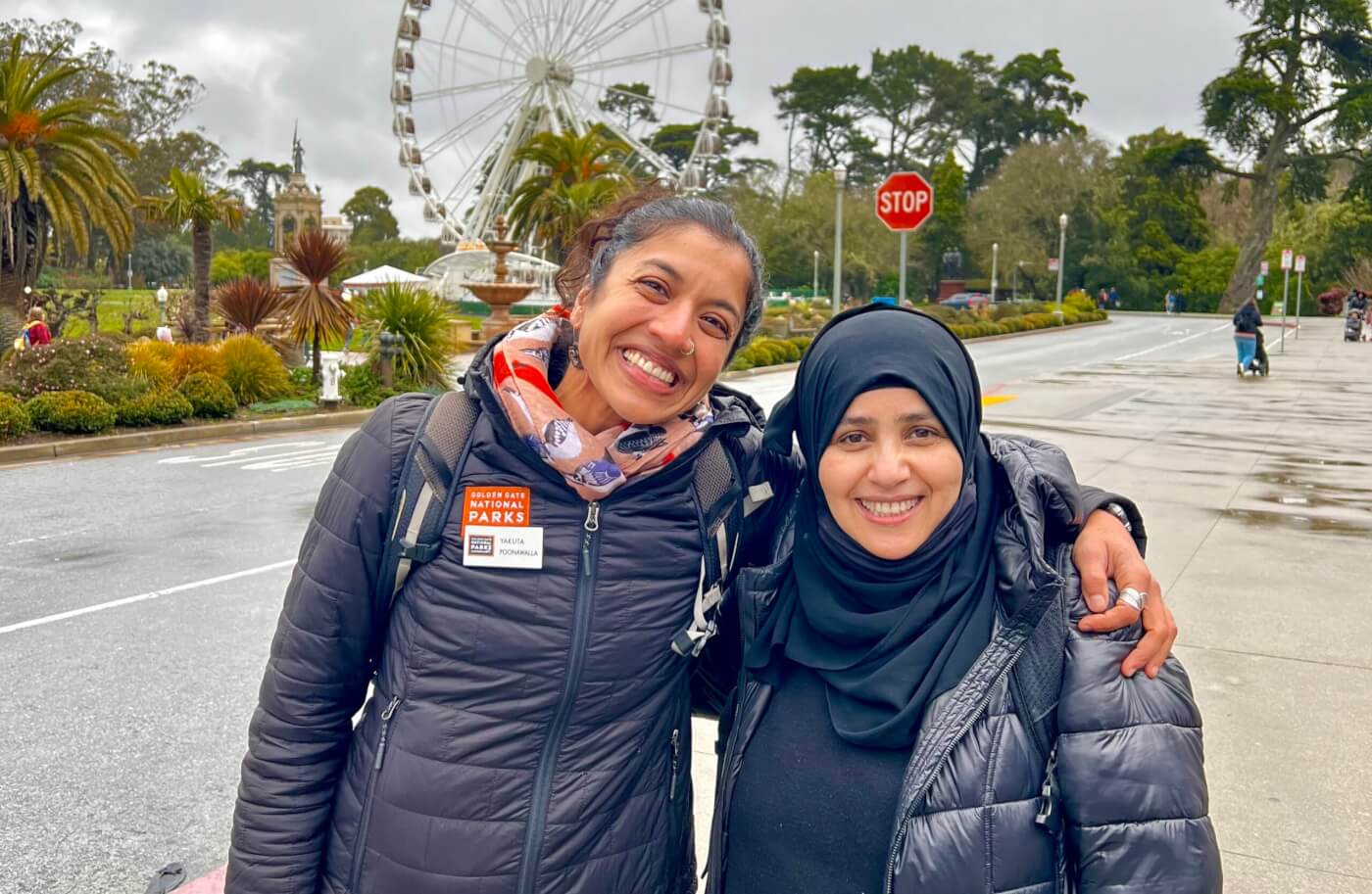 two women smiling at the camera with arms wrapped around each other. There is a ferris wheel and foliage in background.